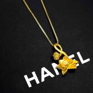 May include: A gold chain necklace with a pendant featuring a stylized lotus flower with a stem and seed pods. The pendant is on a black background with the word "HANEL" printed in white.