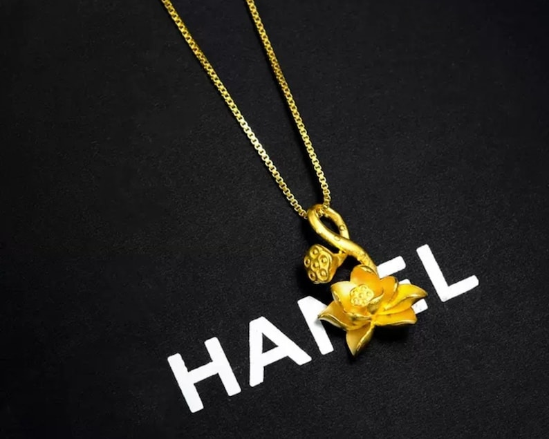 May include: A gold chain necklace with a pendant featuring a stylized lotus flower with a stem and seed pods. The pendant is on a black background with the word "HANEL" printed in white.