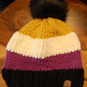 May include: A multi-colored knitted beanie with a black pom pom. The beanie is striped with yellow, white, purple, and black. The beanie has a brown tag with the text "Rainbow Potato Co."