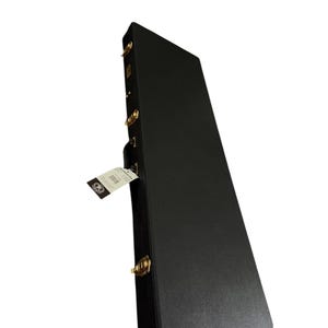 May include: A black rectangular guitar case with gold-colored latches and hinges. A white tag with a logo is attached to the case. The case is angled, showing its side and top.
