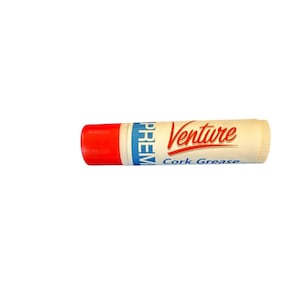 May include: A tube of Venture Cork Grease, with a red cap and white body. The text "Venture" is in red script, and "Cork Grease" is in blue. The tube is designed for musical instrument maintenance.