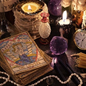May include: A tarot card reading spread with a variety of items including candles, crystals, a pocket watch, and a deck of tarot cards with the "The Lovers" card face up.