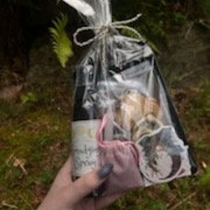 May include: A gift bag filled with various items, including a bottle of wine, a pink ribbon, and a small brown paper bag with a sticker. The bag is tied with twine and wrapped in clear plastic.
