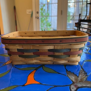 May include: A rectangular woven basket with a green, red, and brown trim. The basket is made of natural wood and has a woven pattern.