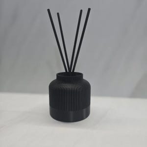 May include: A black reed diffuser. The cylindrical container has a ribbed design and a solid base. Three black reeds are inserted into the container. The background is a light grey.