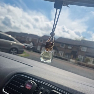 May include: A clear glass bottle with a gold cap hanging from a rear view mirror in a car. The bottle is filled with a clear liquid and has a geometric shape.