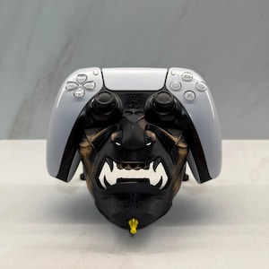 May include: A black and gold decorative face mask with white teeth and a yellow accent, designed to fit a white and black game controller. The controller has buttons and joysticks.
