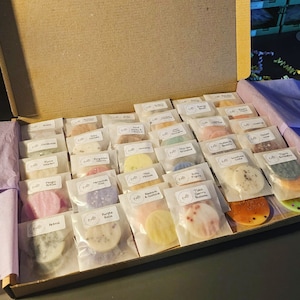 May include: An open box filled with small, round wax melts in various colours and scents. The wax melts are packaged in individual white paper bags with labels that include the scent name. The scents include "Aloha", "Cardamom", "Cocoa Coconut", "Dusk & Tobacco", "Forgotten Cherry", "Magic Potion", "Petrol", "Purple Rain", "Ruby Fig", "Sweet Pea & Cucumber", "Tiast Cherry Blossom", "Vanilla Bean", "Milk & Honey", "Muscat & Blackberry", "Sandalwood Cake", "Forgotten Cherry", "Periwinkle Kiss", "Pink Million", "Floral Sonata", "Lavender & Eucalyptus", "Love Spell", "Coconut Lime", "Beachy Sands", "Energy Boost", "Sweet & Spicy", "Clove & Orange", "Cranberry", "Almond", "Vanilla Bean", "Blackberry & Vanilla", "Dusk & Tobacco", "Cocoa Coconut", "Cardamom", "Aloha".