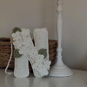 May include: A white crocheted letter "N" decorated with white and green crocheted flowers. The letter is standing on a white surface, with a white candlestick holder in the background. A woven basket is also visible.