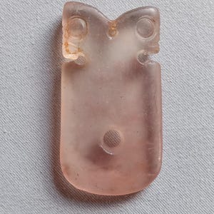 May include: A translucent, pale pink, rectangular pendant with rounded bottom corners. The top features two circular indentations resembling eyes, with a small central hole between them. A larger circular hole is positioned in the lower center.