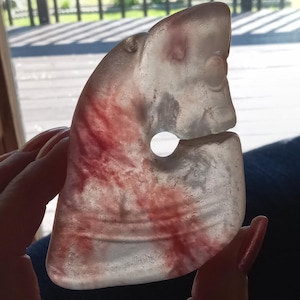 May include: A translucent, semi-circular stone pendant featuring a carved profile of a creature with a circular eye and a central hole. The material is primarily off-white with distinct, irregular streaks of deep red and gray mineral inclusions.