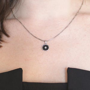 May include: A silver necklace featuring a small, circular pendant. The pendant has a black centre with a white starburst design and a silver frame. The necklace is worn against a person's skin and a black top.