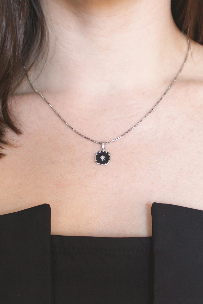 May include: A silver necklace featuring a small, circular pendant. The pendant has a black centre with a white starburst design and a silver frame. The necklace is worn against a person's skin and a black top.