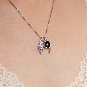 May include: Silver necklace featuring three charms. One charm is a black flower with a white centre, another is a blue and white floral design, and the third is a small clear stone. The necklace is worn against a cream lace top.