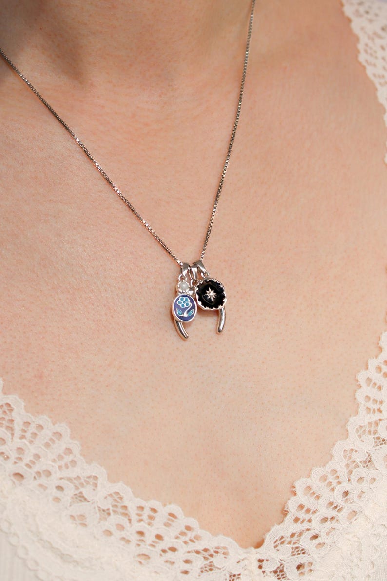 May include: Silver necklace featuring three charms. One charm is a black flower with a white centre, another is a blue and white floral design, and the third is a small clear stone. The necklace is worn against a cream lace top.