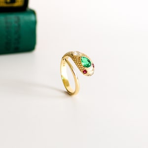 May include: A gold snake ring featuring a textured scale pattern on the head, a pear-shaped green gemstone, a round clear gemstone, and two small red accent stones. The band is smooth and polished, with the text 14K visible on the inner surface.