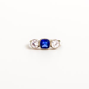 Könnte beinhalten: Ein Ring mit drei Steinen, bestehend aus einem zentralen, kissenförmigen blauen Saphir, der von zwei runden, klaren weißen Steinen flankiert wird. Die Steine sind in eine silberfarbene Ringschiene mit feiner Perlenstruktur eingefasst.