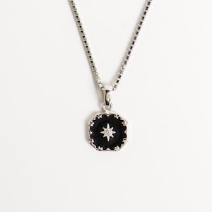 May include: Silver necklace featuring a black pendant with a scalloped edge and a starburst design, with a small, clear gem at its centre. The chain is a box style.