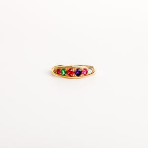 Op de afbeelding: Een gouden ring met een rij van vijf kleine, ronde, veelkleurige edelstenen. De stenen zijn gerangschikt in een volgorde van roze, groen, rood, paars en een heldere witte steen, horizontaal gezet in de gepolijste gouden metalen band.