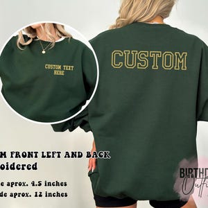 May include: A dark green sweatshirt with the word "CUSTOM" embroidered in gold on the back. The front of the sweatshirt has a smaller gold embroidered design that says "CUSTOM TEXT HERE". The sweatshirt is 4.5 inches wide on the left side and 12 inches wide on the back.
