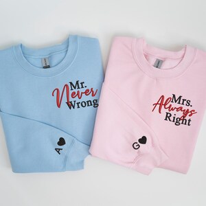 May include: Two sweatshirts, one light blue and one pink, with embroidered text. The blue sweatshirt says "Mr. Never Wrong" and the pink sweatshirt says "Mrs. Always Right". Both sweatshirts have a small black heart with a letter embroidered on the sleeve.