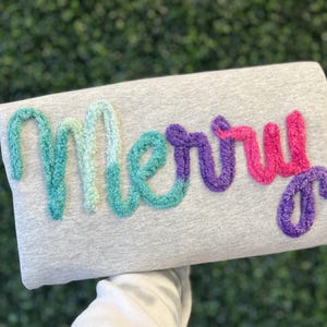 May include: A gray sweatshirt with the word "Merry" embroidered in colorful, textured yarn. The word "Merry" is in shades of teal, purple, and pink, creating a festive and eye-catching design.