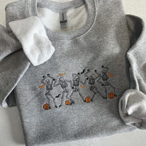 May include: A heather grey crewneck sweatshirt featuring an embroidered design of five dancing skeletons. The skeletons are positioned above small orange pumpkins and are surrounded by tiny black bats and orange leaves, creating a festive scene.