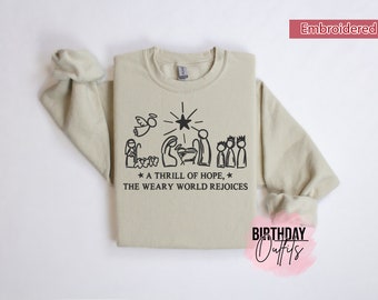 Op de afbeelding: Een beige sweatshirt met een zwart geborduurd ontwerp van een kerststal met de tekst "* A THRILL OF HOPE, * THE WEARY WORLD REJOICES".