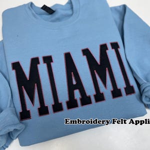 May include: A light blue sweatshirt with the word "MIAMI" in large, black, block letters with pink stitching. The text "Embroidery Felt Applique" is printed below the word "MIAMI".