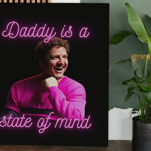 Peut inclure: Impression sur toile encadrée avec l'inscription "Daddy is a state of mind" en néon rose. L'image présente un homme en pull rose, souriant et riant. Le fond est noir.