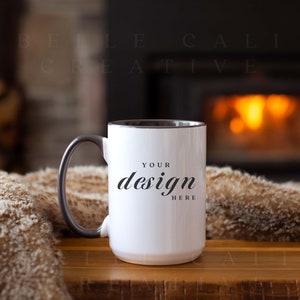 May include: White ceramic mug with a black rim and handle. The mug has the text "Your design here" printed on the front. The mug is sitting on a wooden table in front of a fireplace.