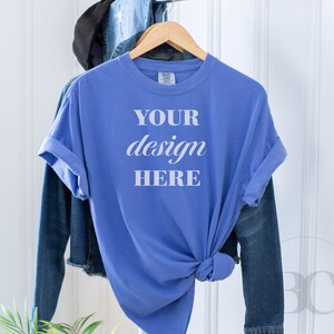 May include: A blue t-shirt with the text "YOUR design HERE" on a hanger. The t-shirt is hanging in front of a white wall with a denim jacket draped over the hanger.
