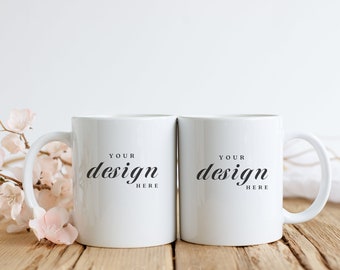 Puede incluir: Dos tazas de cerámica blanca con texto negro que dice "Your design here" sobre una superficie de madera con flores rosas.