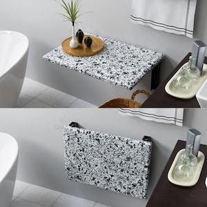 May include: A white and black speckled folding wall table with a black metal frame. The table is mounted to a wall and is folded up against the wall. The table is in a bathroom setting.