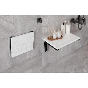 May include: White folding wall mounted shelf with black metal brackets. The shelf is mounted on a tiled wall and has a wooden top. There are three bottles of liquid soap on the shelf.
