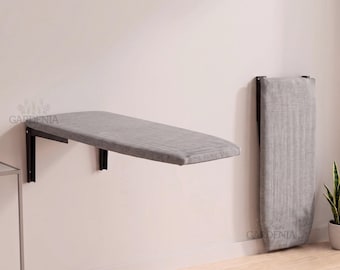 May include: A wall-mounted ironing board with a grey fabric cover and black metal support brackets. The board is shown in two positions: one extended horizontally for use and one folded vertically against the wall for storage and space saving.