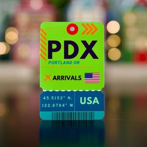 May include: Green and blue airport arrival tag with the text "PDX" and "PORTLAND OR" in white. The tag also has the text "ARRIVALS" and a small American flag. The coordinates "45.5152° N, 122.6784° W" and "USA" are printed on the tag.