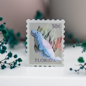 May include: A blue and green watercolor illustration of a manatee on a vintage-style postage stamp. The stamp is labeled "50&cent;" and "FLORIDA".