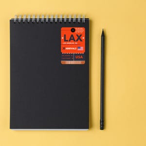 May include: A black notebook with a spiral binding and a bright orange luggage tag sticker that says "LAX Los Angeles, CA Arrivals 34.0548&deg; N 118.2428&deg; W USA".