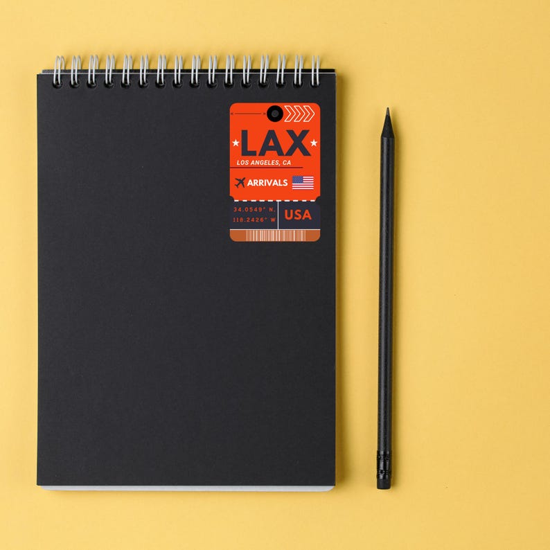 May include: A black notebook with a spiral binding and a bright orange luggage tag sticker that says "LAX Los Angeles, CA Arrivals 34.0548&deg; N 118.2428&deg; W USA".