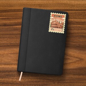 May include: Black leather-bound journal with a Rome-themed postage stamp sticker. The stamp features an illustration of the Colosseum, a scooter, and the number 5. A peach-colored ribbon bookmark is visible.