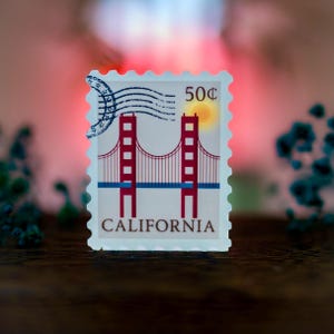 Pode incluir: Um selo postal vintage de 50 c&ecirc;ntimos com uma ilustra&ccedil;&atilde;o vermelha e branca da Golden Gate Bridge em S&atilde;o Francisco, Calif&oacute;rnia.