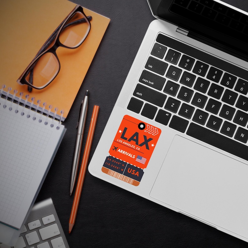 May include: Orange and black airport luggage tag sticker with the text "LAX" and "Los Angeles, CA" and "Arrivals" with a small American flag. The sticker is on a silver laptop.