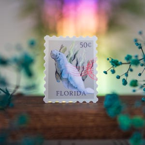 May include: A vintage-style Florida postage stamp featuring a watercolor illustration of a manatee swimming in a bed of seaweed. The stamp is labeled "50&cent;" and "FLORIDA".
