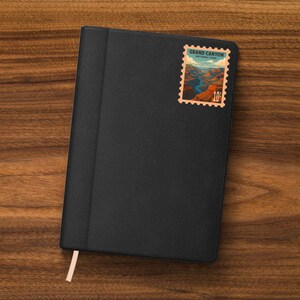 K&ouml;nnte beinhalten: Schwarzes Lederjournal mit einem Lesezeichen. Eine Vintage-Briefmarke des Grand Canyon National Park ist in der oberen rechten Ecke angebracht. Die Briefmarke zeigt eine Landschaftsansicht des Canyons und den Text "Grand Canyon National Park" und "10&cent;".