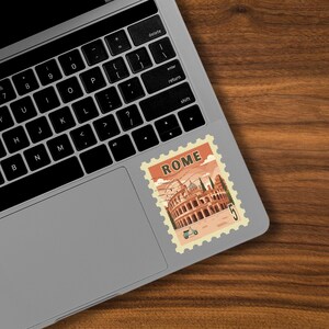 May include: A sticker featuring a vintage-style illustration of the Colosseum in Rome, Italy, with the word "ROME" at the top. The sticker has a postage stamp design and is placed on a laptop.
