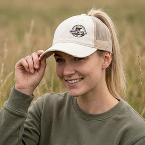 May include: A cream-colored baseball cap with a distressed brim and a tan mesh back. The front features a circular black graphic with a silhouette of a cow and the text DROSTE CATTLE CO. SIOUX CITY, IA.
