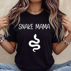 Könnte beinhalten: Schwarzes T-Shirt mit dem weißen Schriftzug "SNAKE MAMA" und einer weißen Schlangenillustration. Das Shirt hat einen Rundhalsausschnitt.