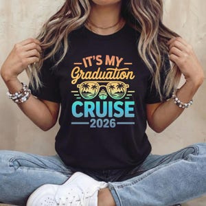 May include: Black t-shirt with the text "IT'S MY Graduation CRUISE 2026" in a gradient of yellow, orange, and blue. The design includes sunglasses with palm trees and a cruise ship. The shirt is worn by a person.