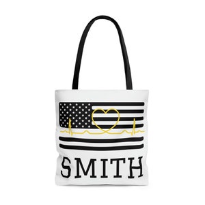 May include: White tote bag with a black and white American flag design. A gold heart and EKG line are superimposed on the flag. The text "SMITH" is printed in black below the flag.